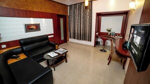 A hotel lobby with a black leather sofa, a small wooden table with a magazine, a TV screen displaying a movie poster, and a reception desk with a mirror and a vase of flowers. - Hotel RKV Residency (Krishnagiri)