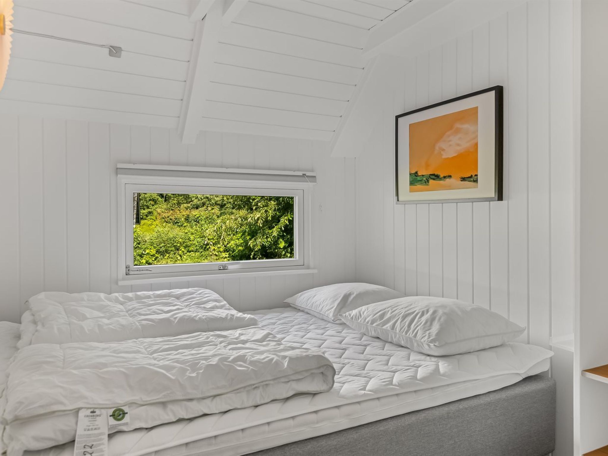 A bedroom with a bed, a window with a view of greenery, and a framed picture on the wall.