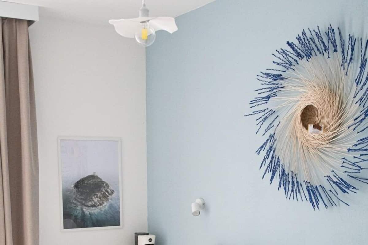 Ein ordentlich bezogenes Bett mit weißen und grauen Kissen, ein weißer Schreibtisch mit Kaffeemaschine, eine Wandleuchte und ein gerahmtes Bild an der Wand.