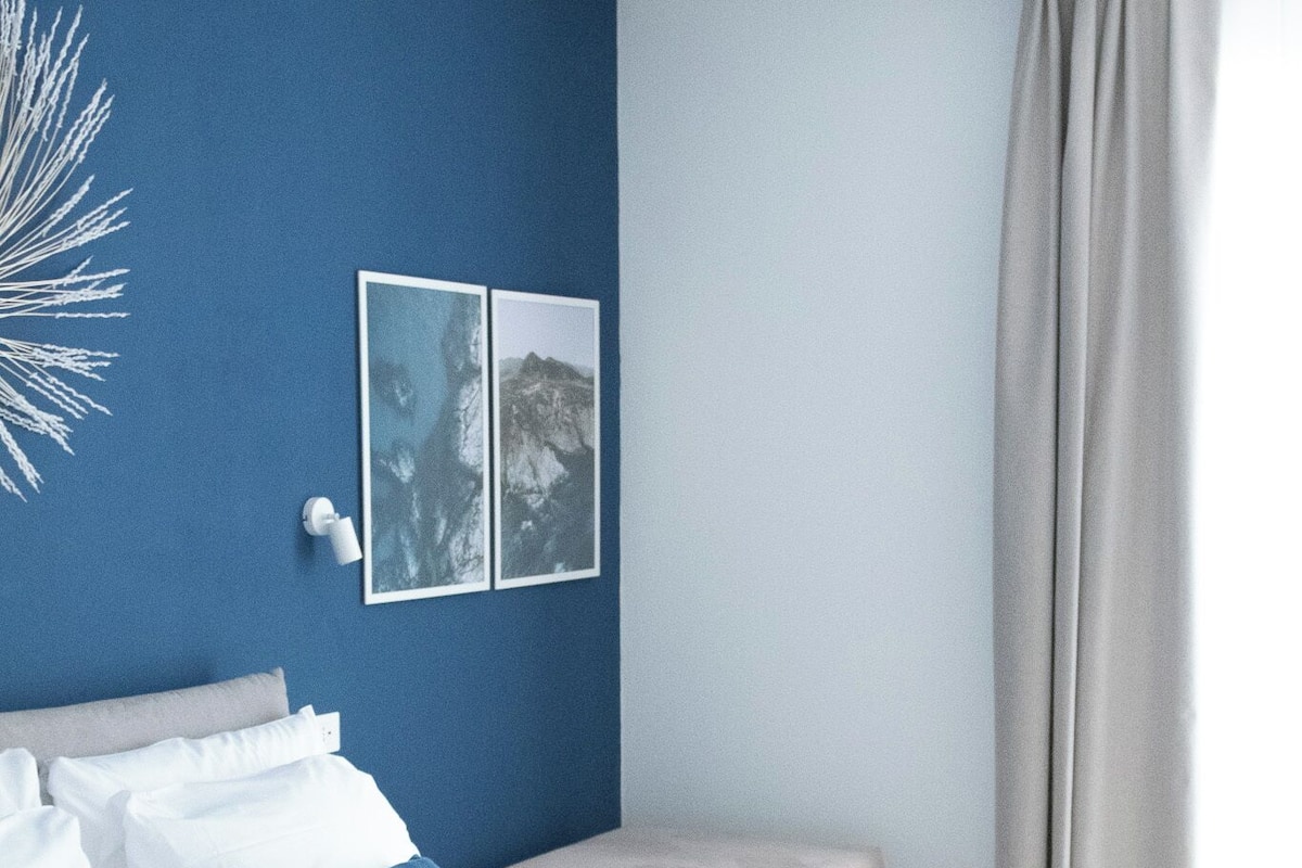Ein ordentlich bezogenes Bett mit weißen Leinen und blauen Kissen, ein Nachttisch mit Lampe sowie eine große Wanddekoration.