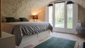 Un dormitorio con techo de piedra, cama con ropa de cama gris, alfombra verde y mesita de noche con lámpara.