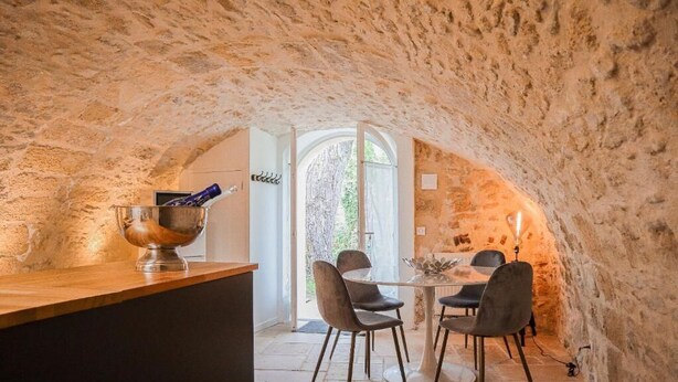 Interior rústico de piedra con una mesa redonda para comer y sillas, un cuenco sobre una encimera de madera, y una puerta que da al exterior.