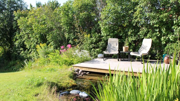 A wooden deck with two chairs and a table, surrounded by lush greenery and a pond. - Cozy and spacious traditional Swedish Dala cottage (Dalarnas län)