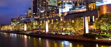 A waterfront hotel with modern architecture, a river in the foreground, and illuminated windows.