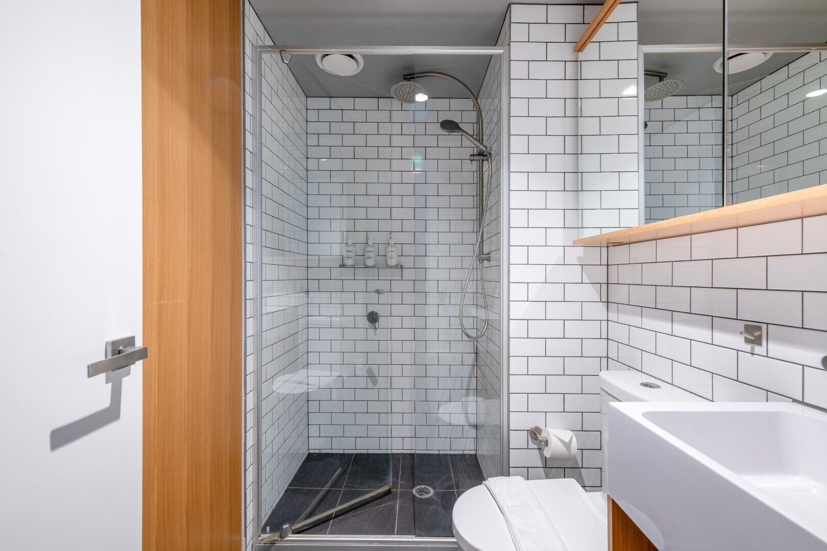 Une salle de bain moderne avec des carreaux de céramique blancs rectangulaires, une douche en verre, un lavabo blanc et un meuble-lavabo en bois.