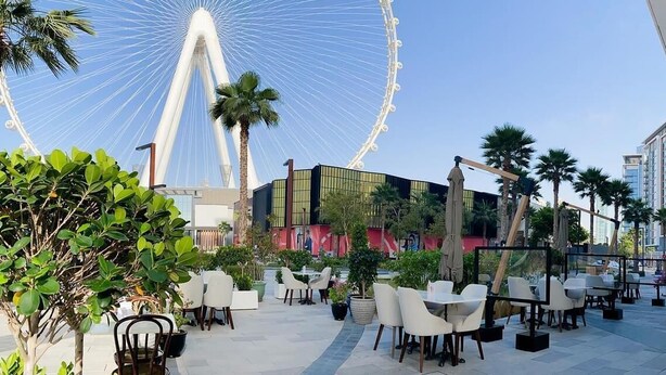 Ein Café mit Sitzgelegenheiten im Freien, im Hintergrund ein Riesenrad und Palmen.