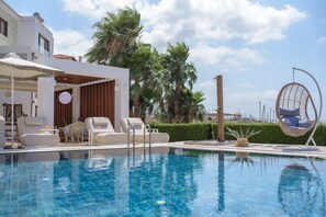 Una piscina con sillas de descanso y una hamaca colgante, rodeada de palmeras y un edificio de fachada blanca.