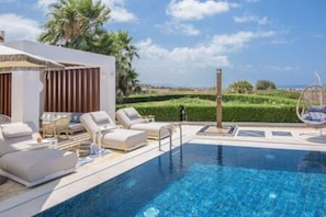 Un área de piscina moderna con sillones de descanso y una hamaca colgante, rodeada de follaje y un cielo despejado.