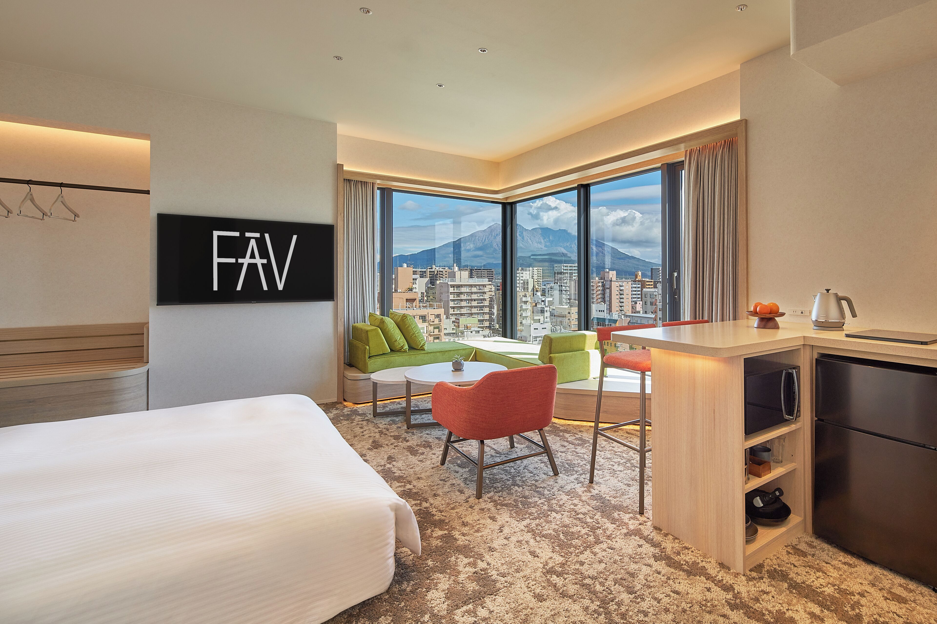 A hotel room with a bed, a desk, a chair, a sofa, a large window with a city and mountain view, and a minibar.
