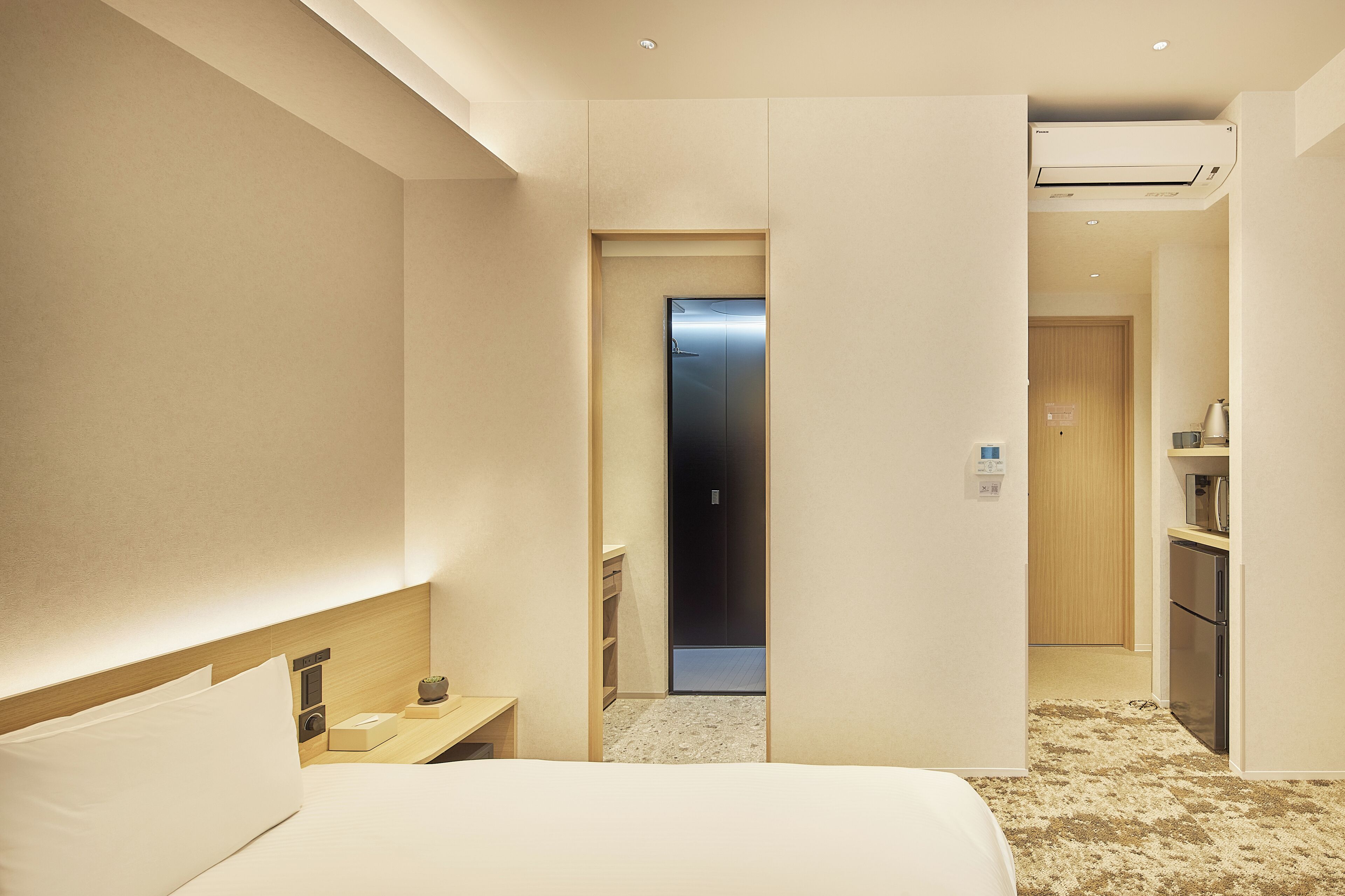 A hotel room with a bed, bedside table, and a small kitchenette area.