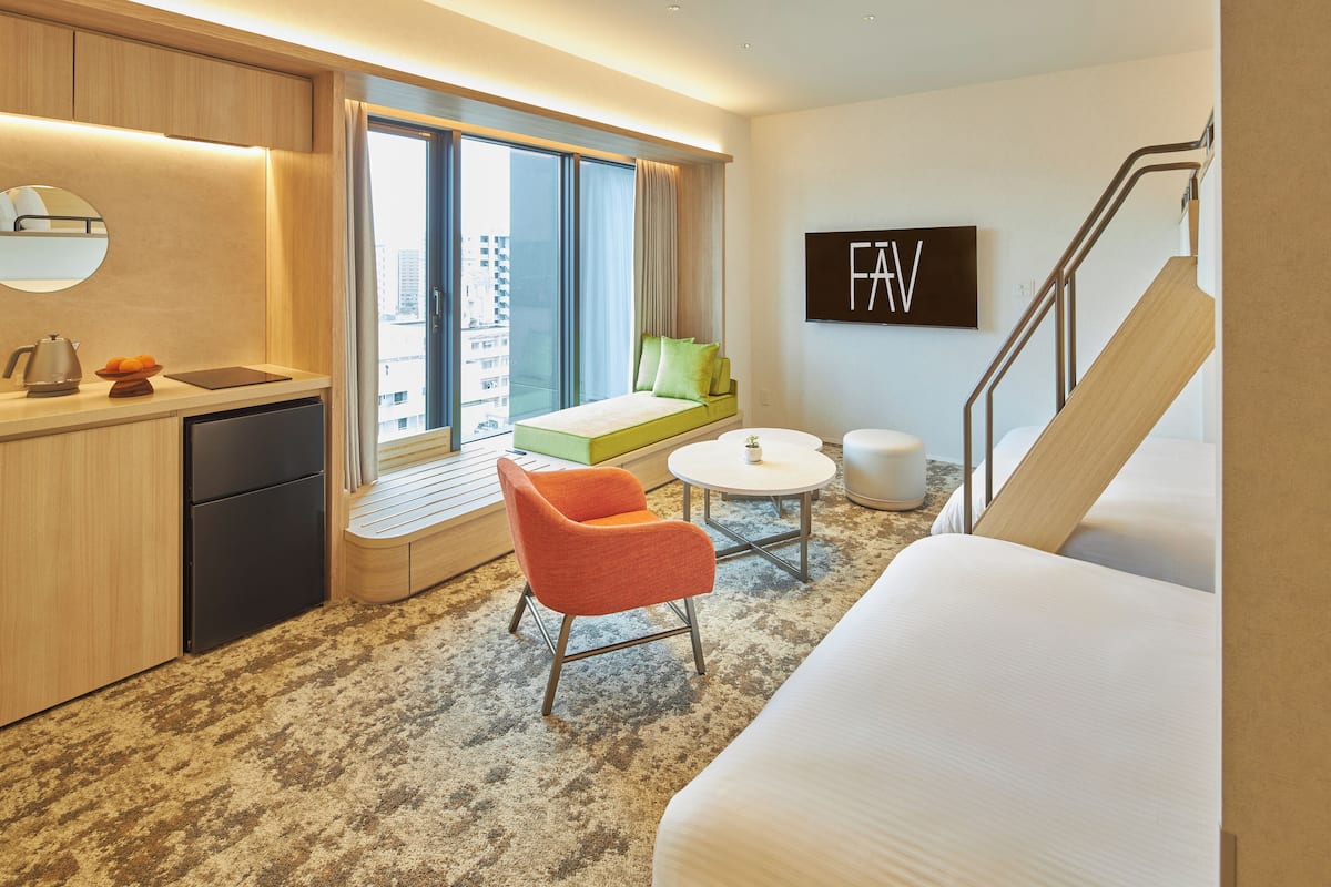 A modern hotel room with a bed, a seating area with a green sofa and orange chair, a small round table, and a view of the city through large windows.
