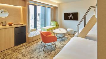A modern hotel room with a bed, a seating area with a green sofa and orange chair, a small round table, and a view of the city through large windows.