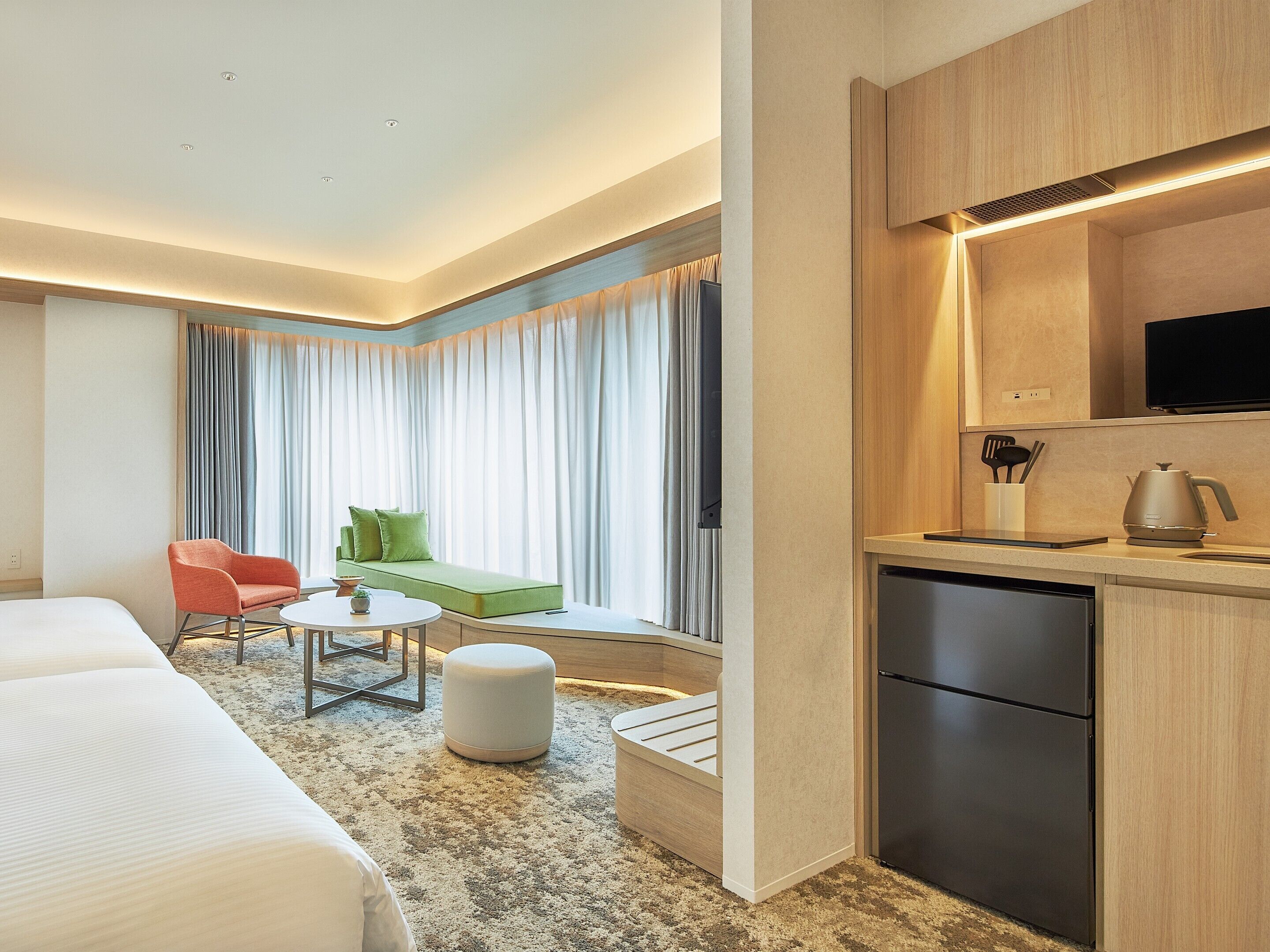 Une chambre d’hôtel avec un lit, un canapé vert, une chaise rouge, une petite table et un coin cuisine équipé d’un réfrigérateur et d’une bouilloire.