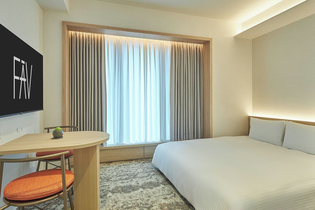 A hotel room with a bed, a desk with a chair, a small plant, and a window with curtains.
