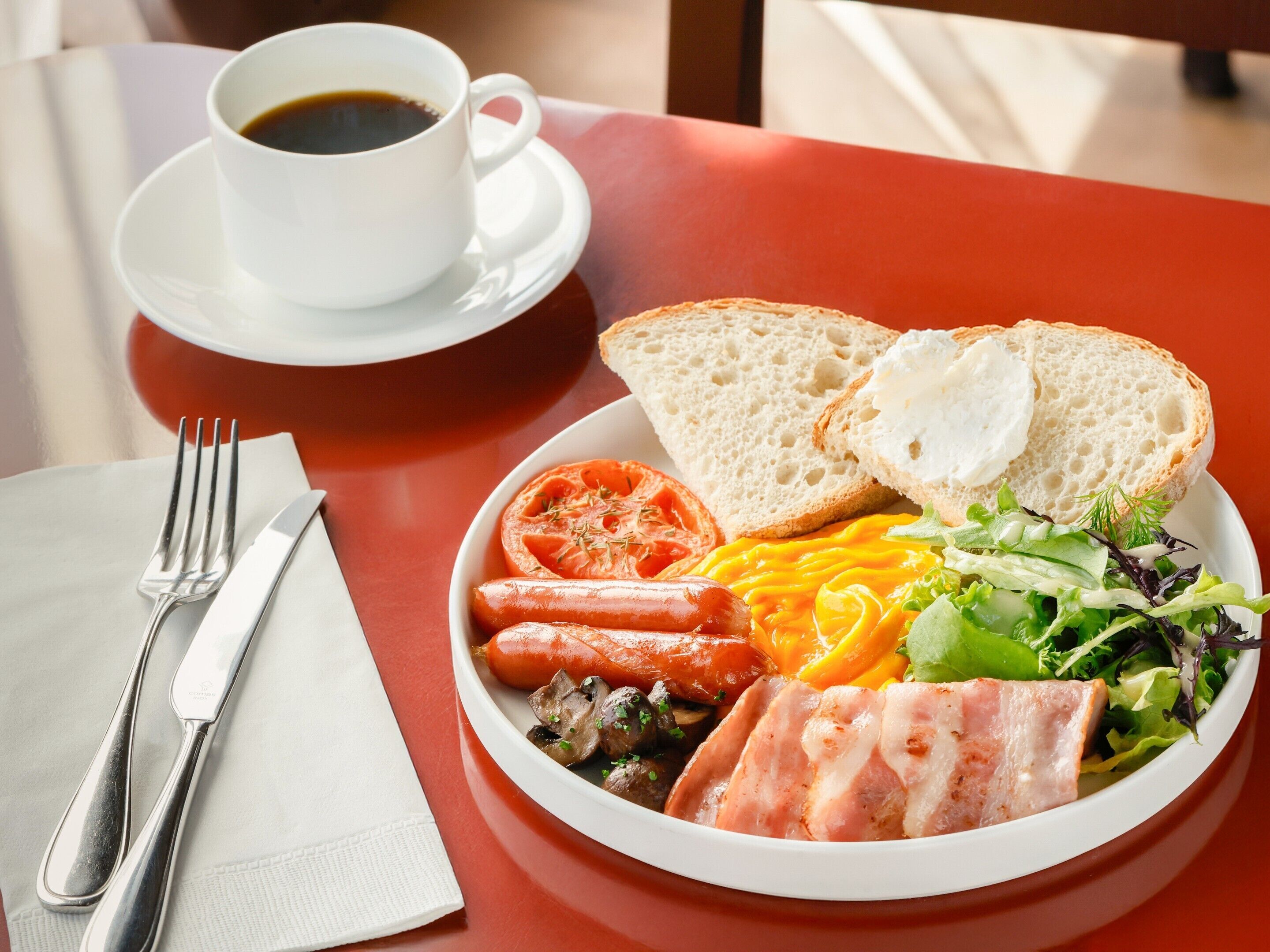 Een bord eten met brood, tomaat, kaas, ham, champignons en salade, een kop koffie, en bestek op een tafel.