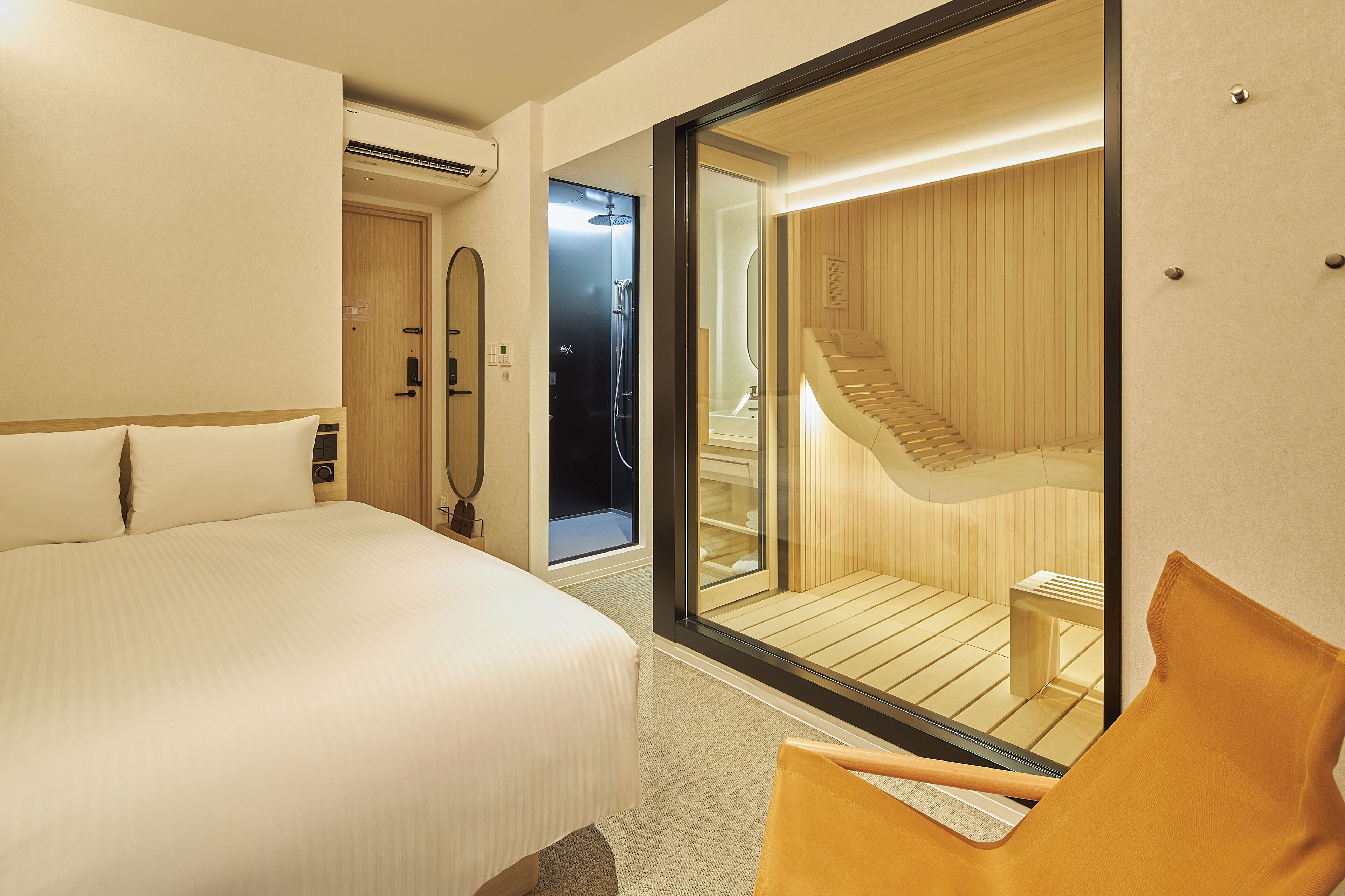 A hotel room with a large bed, a wooden chair, a glass wall, and a visible bathroom with a shower and a wooden staircase.