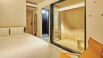 A hotel room with a large bed, a wooden chair, a glass wall, and a visible bathroom with a shower and a wooden staircase.