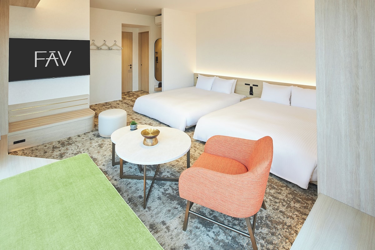 A hotel room with two beds, a small table with a bowl, a green rug, and an orange chair.