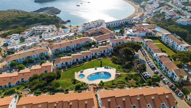 Un complex turístic a la costa amb diversos edificis, una piscina central i vistes al mar.