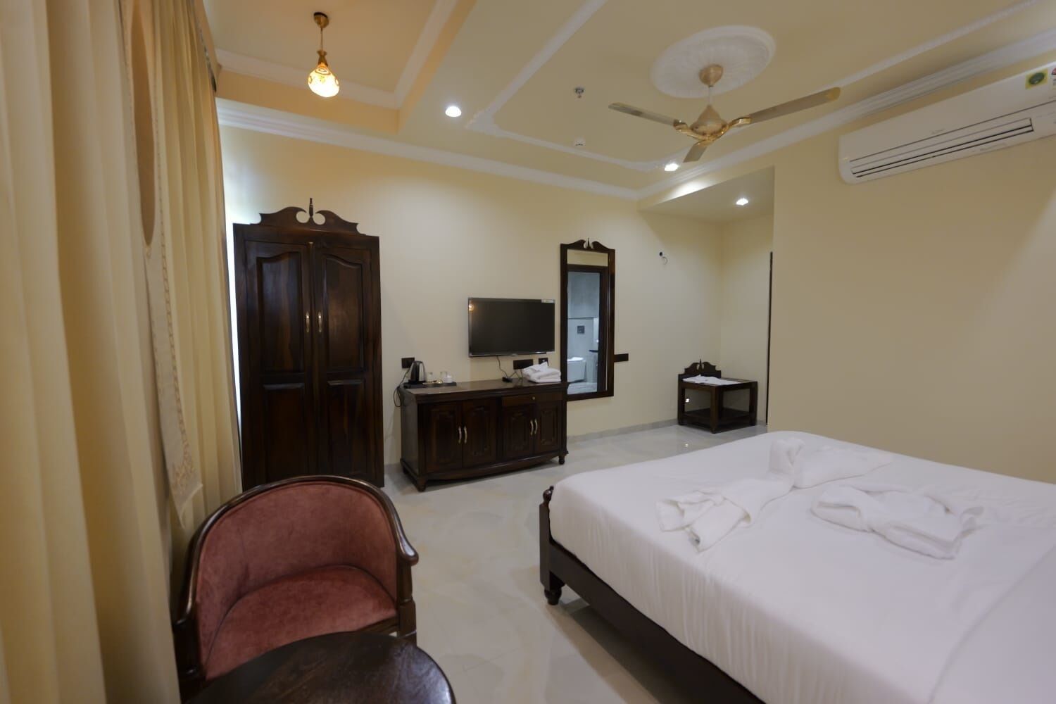A hotel room with a bed, a TV, a wardrobe, a mirror, a ceiling fan, and an air conditioning unit.