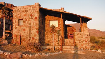 Edificio de piedra con puerta y ventana de madera, rodeado de vegetación desértica.