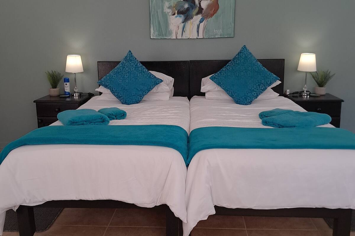 Un lit double avec une literie aux couleurs turquoise et blanche, deux tables de chevet avec des lampes, un tableau au mur et un sol carrelé.