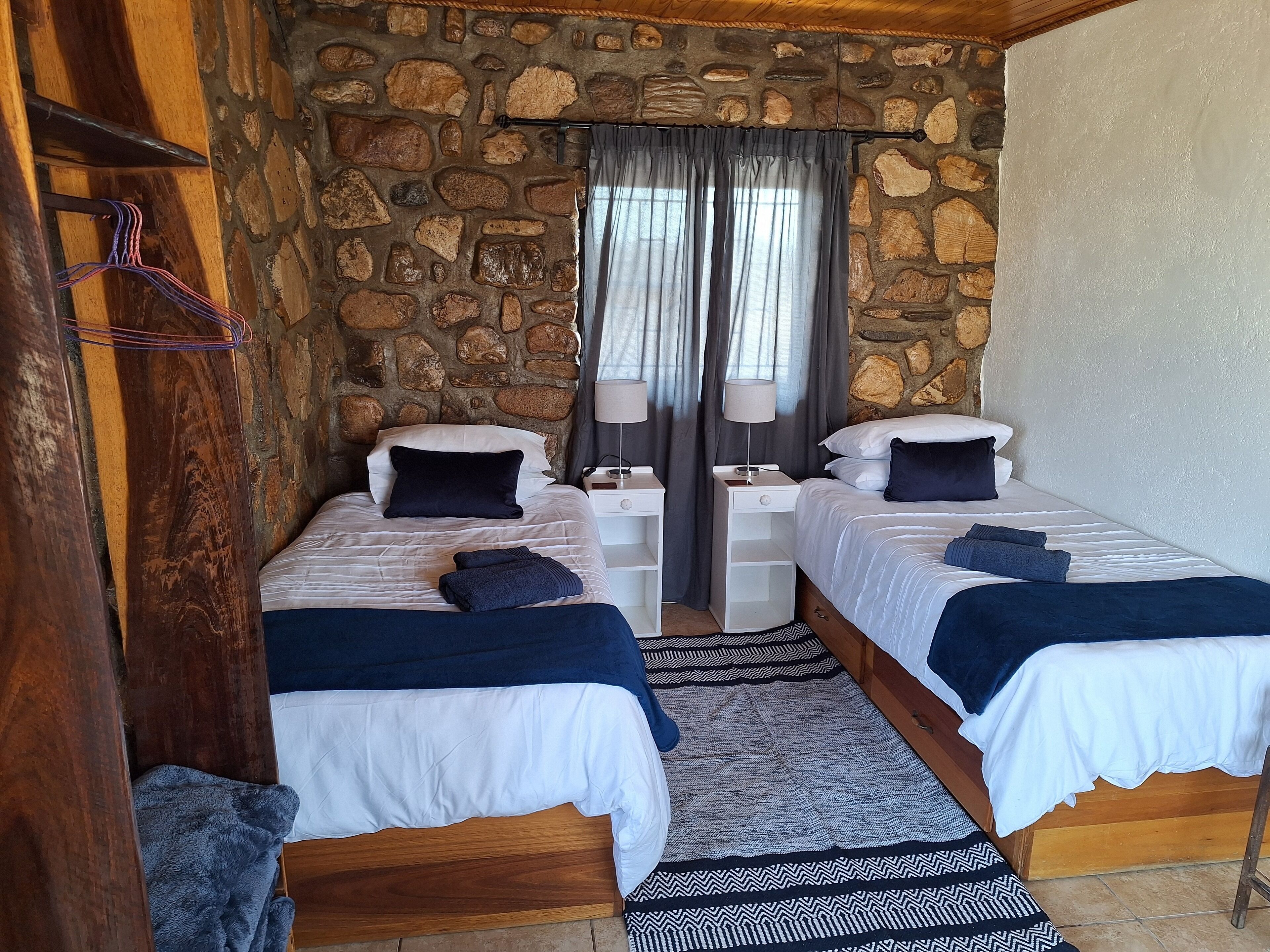 A room with two beds, a stone wall, a wooden ceiling, and a window with curtains.