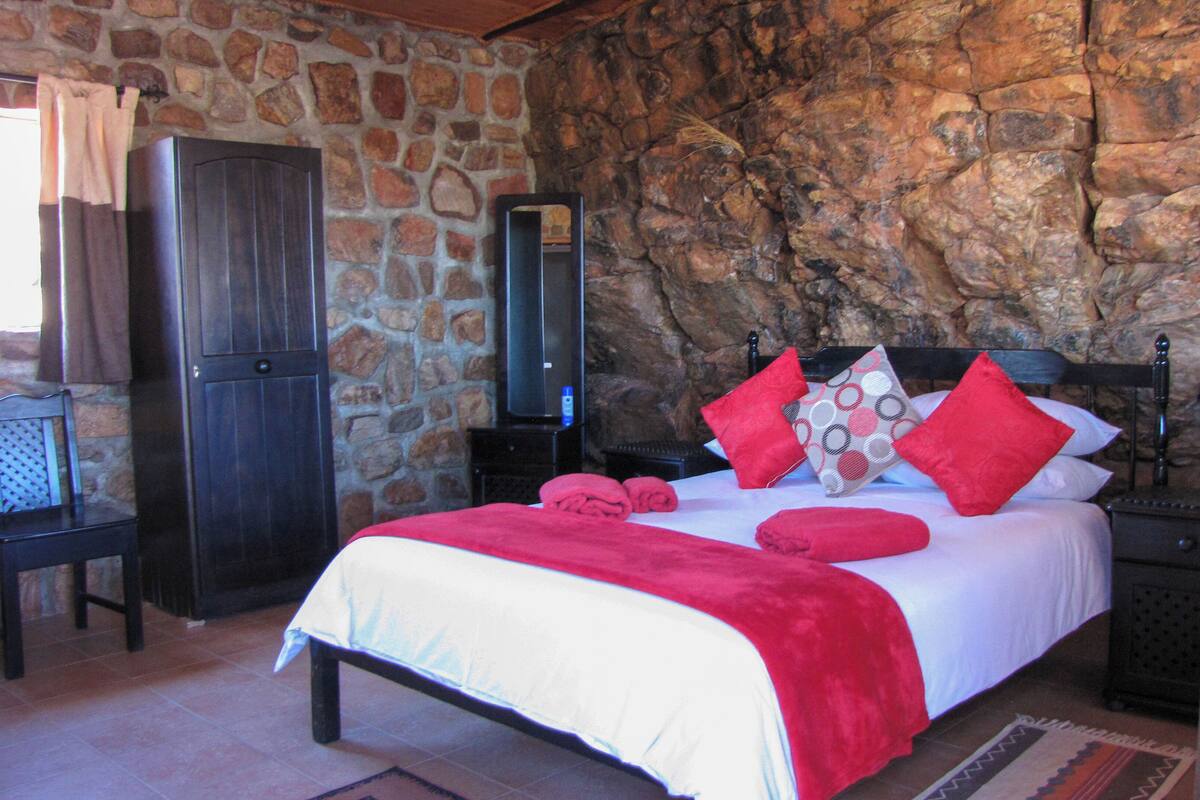 Une chambre avec un mur en pierre, un lit avec une literie rouge, une armoire en bois et une table de chevet avec une lampe.