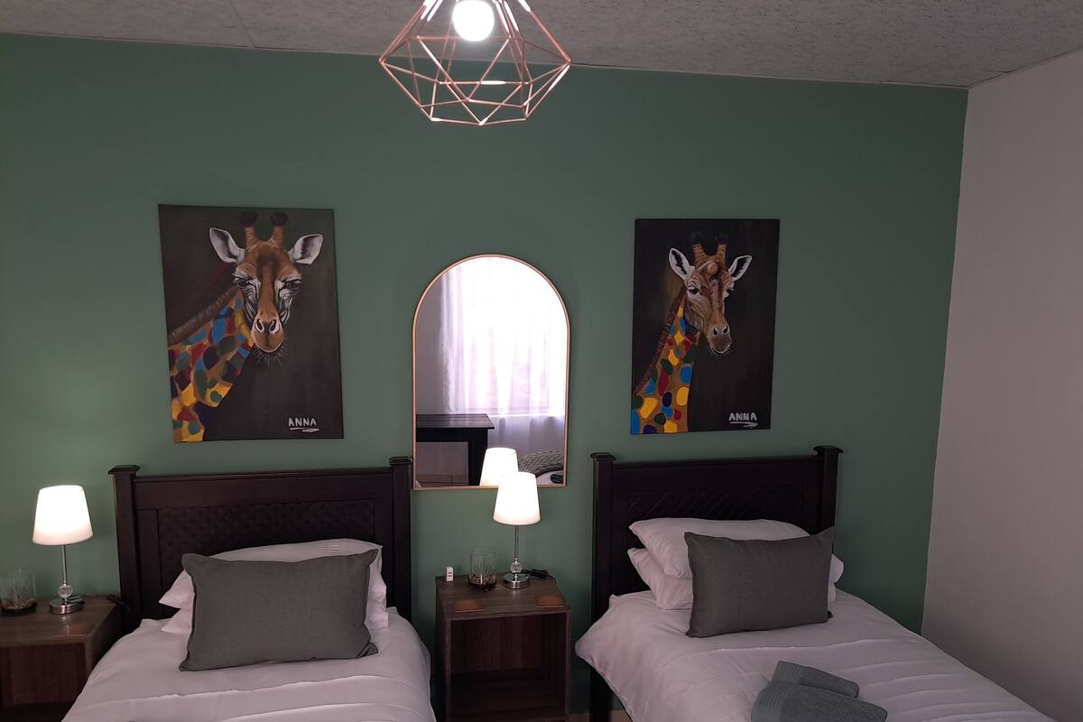 Une chambre d’hôtel avec deux lits, chacun étant équipé d’un oreiller et d’une couverture. On y trouve deux tableaux représentant des girafes, accrochés au mur, un miroir et un luminaire au design géométrique.
