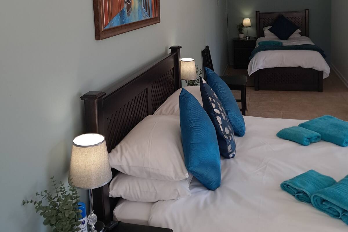 Une chambre d’hôtel avec un grand lit, un lit plus petit, un tableau représentant un paysage urbain, une table de chevet avec une lampe et une plante en pot.