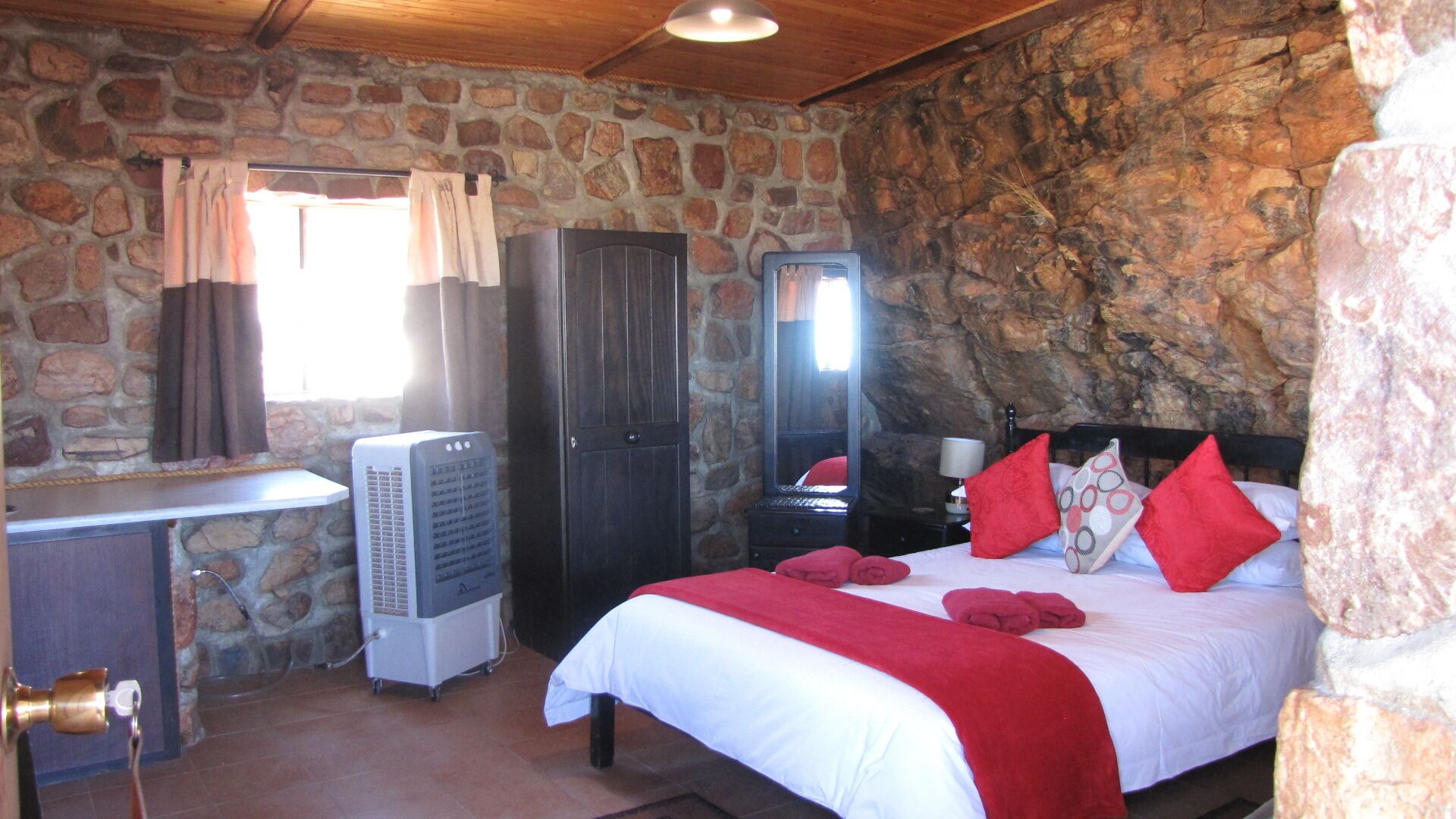 Una habitación con pared de piedra, cama con almohadas rojas, armario, espejo y un calentador.