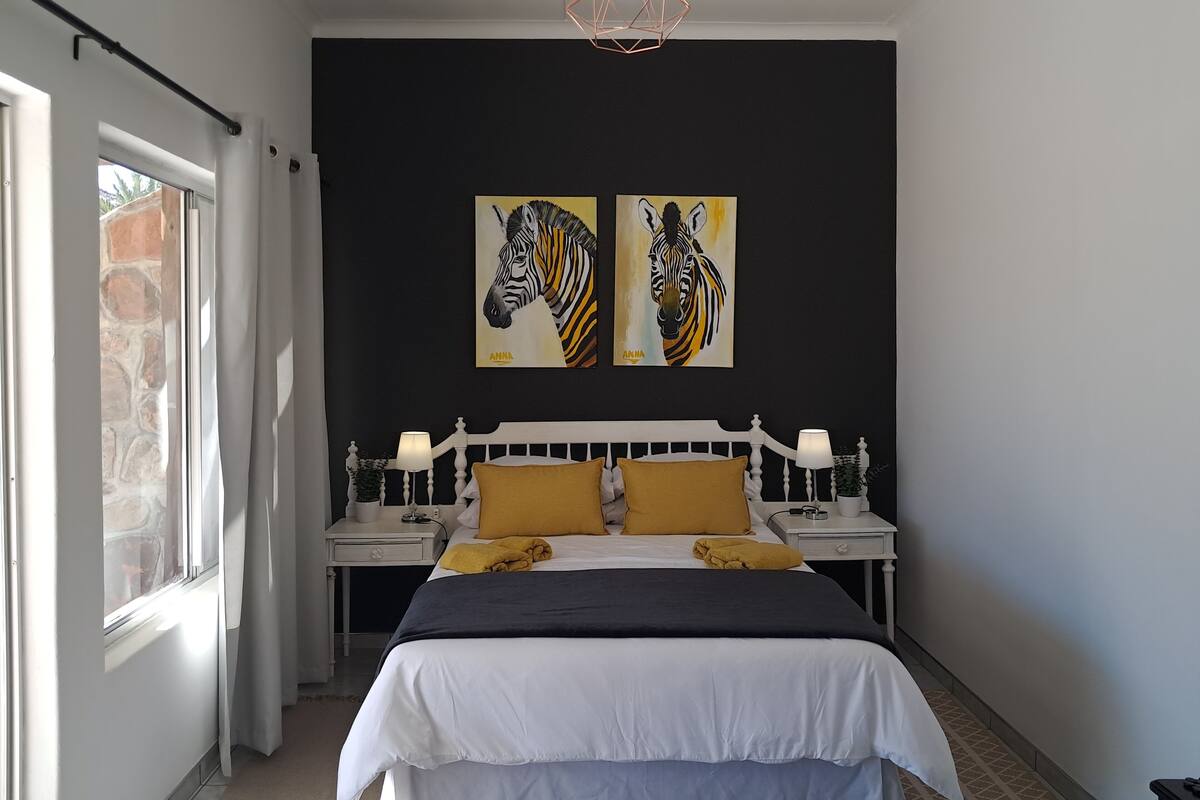 Une chambre à coucher avec un lit, deux lampes fixées au mur, une fenêtre et deux tableaux représentant des zèbres.
