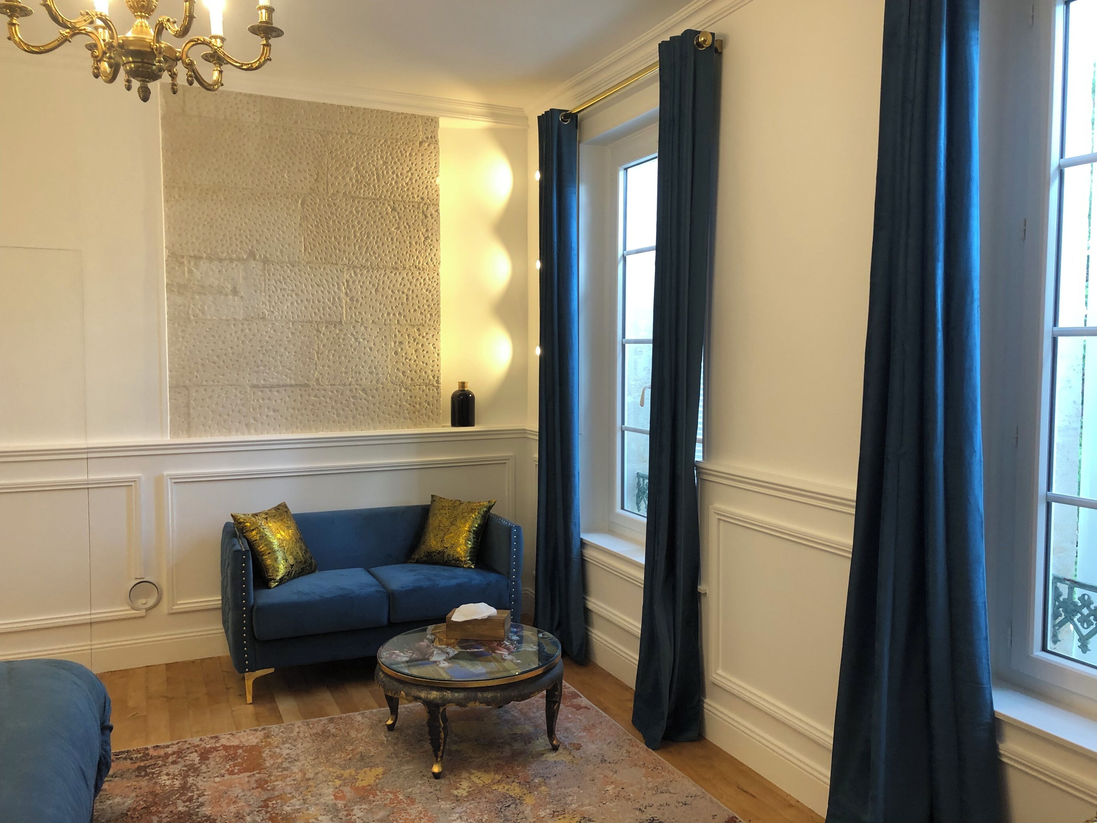 Una habitación con sofá azul, una mesita de centro redonda de cristal, pared con textura y una ventana con cortinas oscuras.