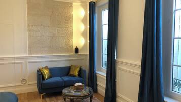 Una habitación con sofá azul, una mesita de centro redonda de cristal, pared con textura y una ventana con cortinas oscuras.