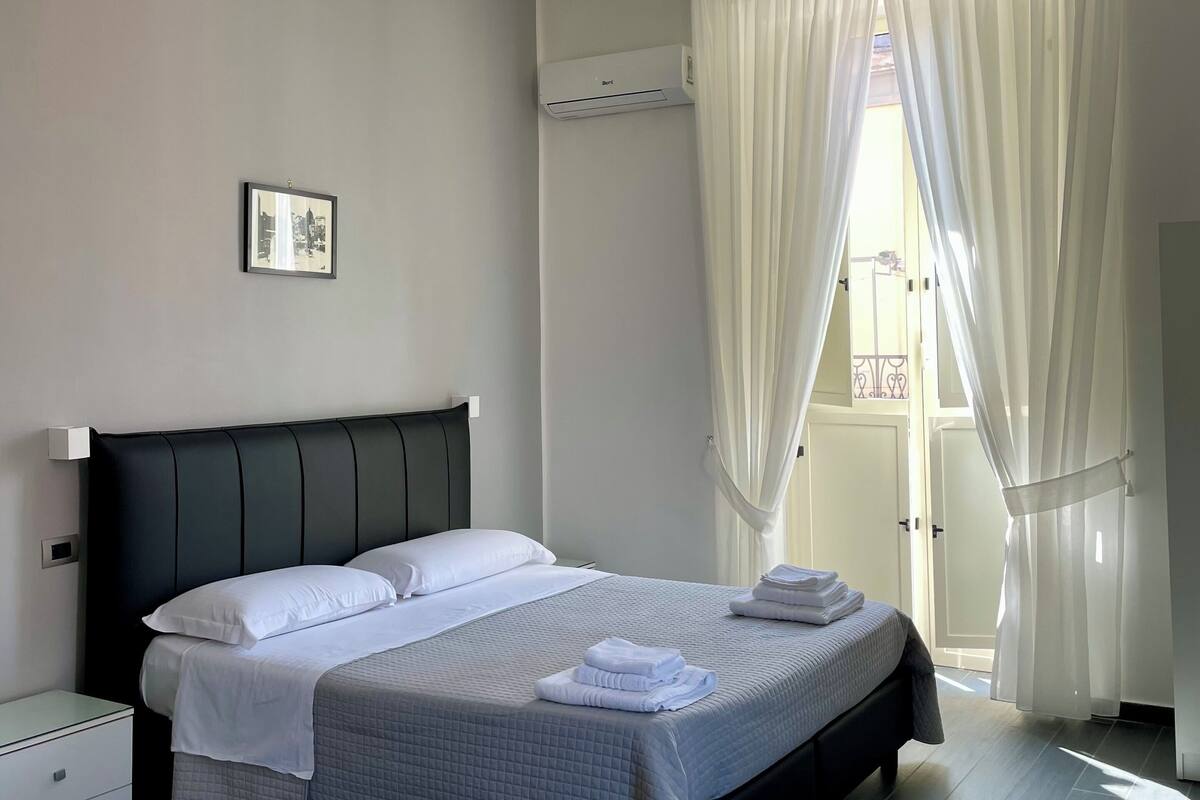 A modern bedroom with a large bed, a bedside table, an air conditioning unit, and a window with sheer curtains.