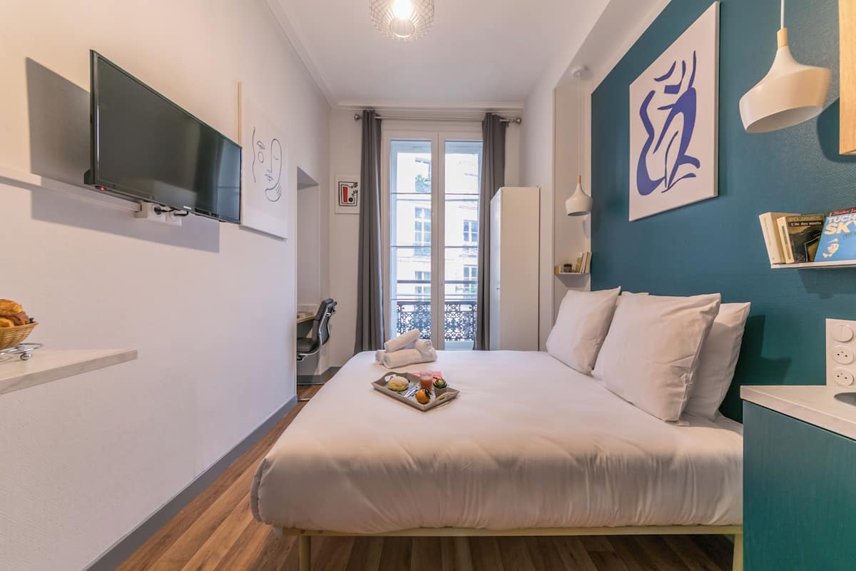 Une chambre d’hôtel avec un lit, un téléviseur à écran plat fixé au mur, une fenêtre avec des rideaux, une petite table avec un plateau de nourriture, et un mur d’accent bleu orné d’un grand tableau abstrait.