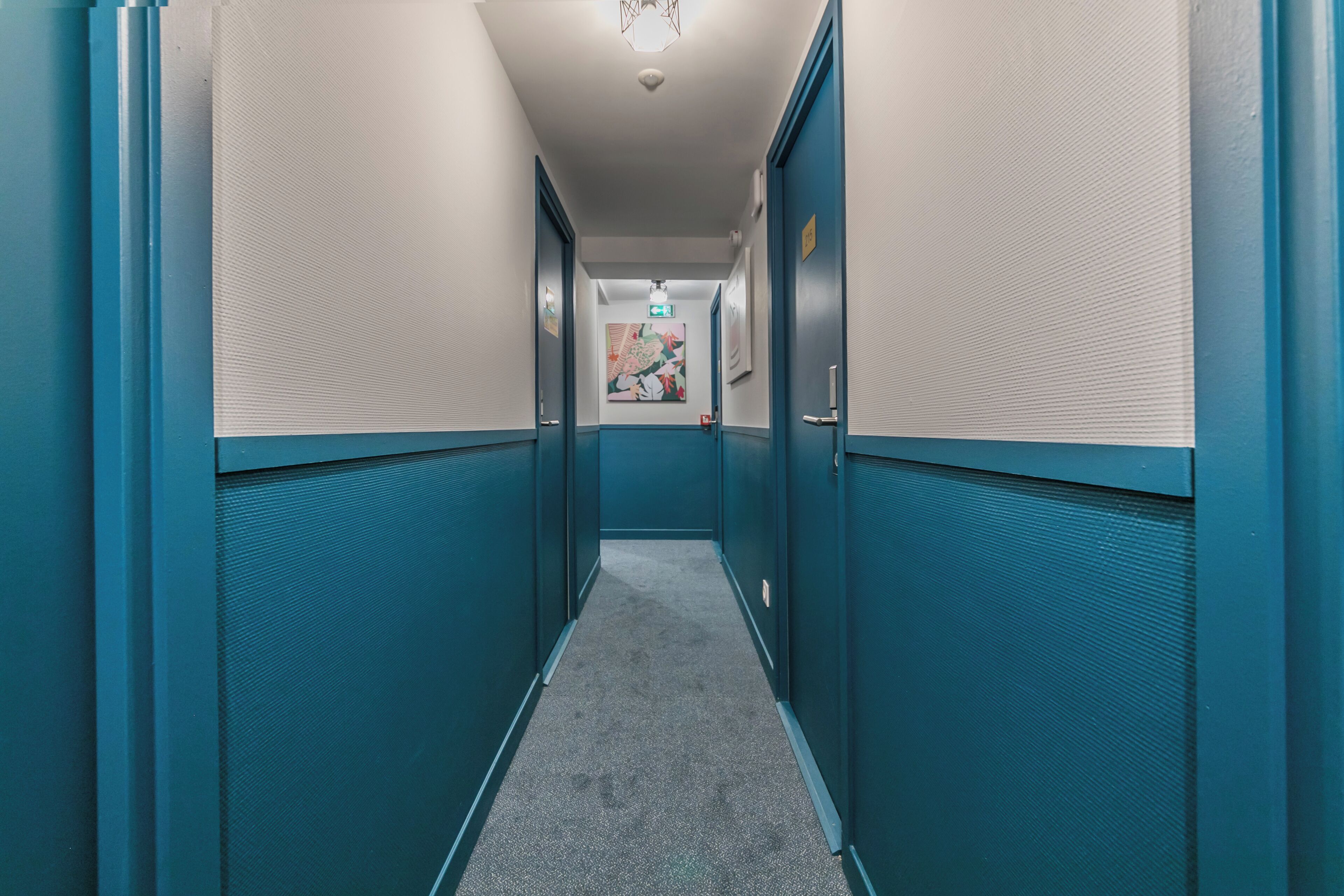 Un pasillo estrecho con paredes color turquesa y suelo alfombrado, que conduce a una puerta con una obra de arte colorida en la pared.