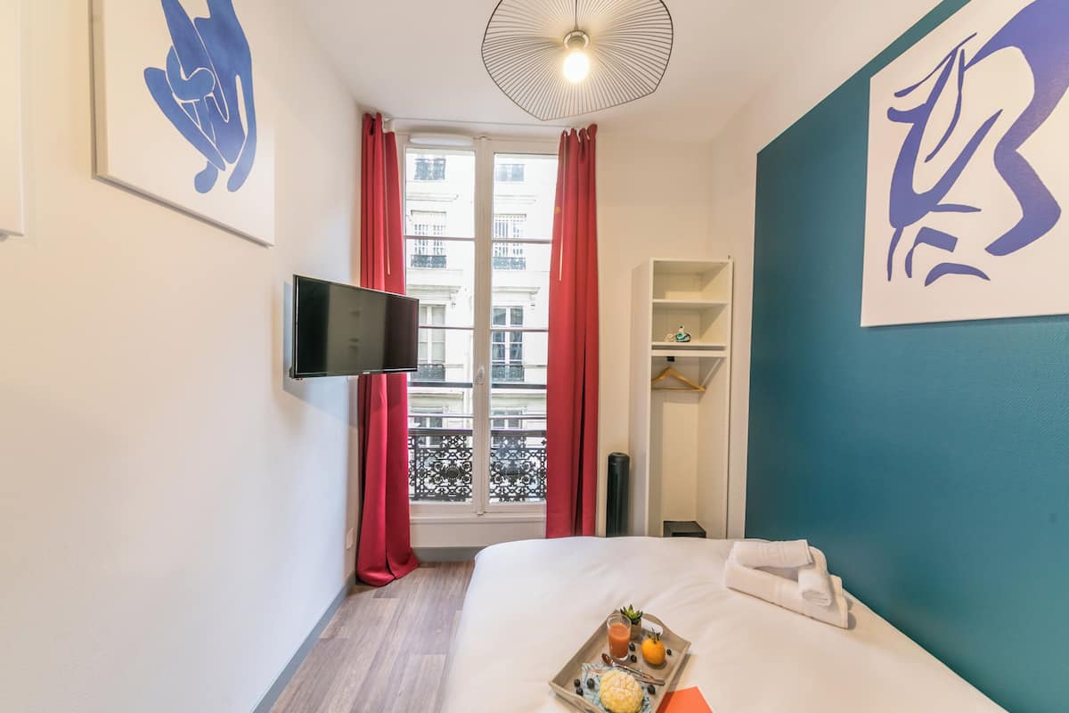 Une chambre d’hôtel avec un lit, un téléviseur fixé au mur, une fenêtre avec des rideaux rouges et des œuvres d’art accrochées aux murs.