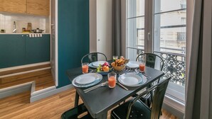 A dining area with a table set for two, a window with a view of buildings, and a kitchen in the background. - WS Louvre - Richelieu (Paris)