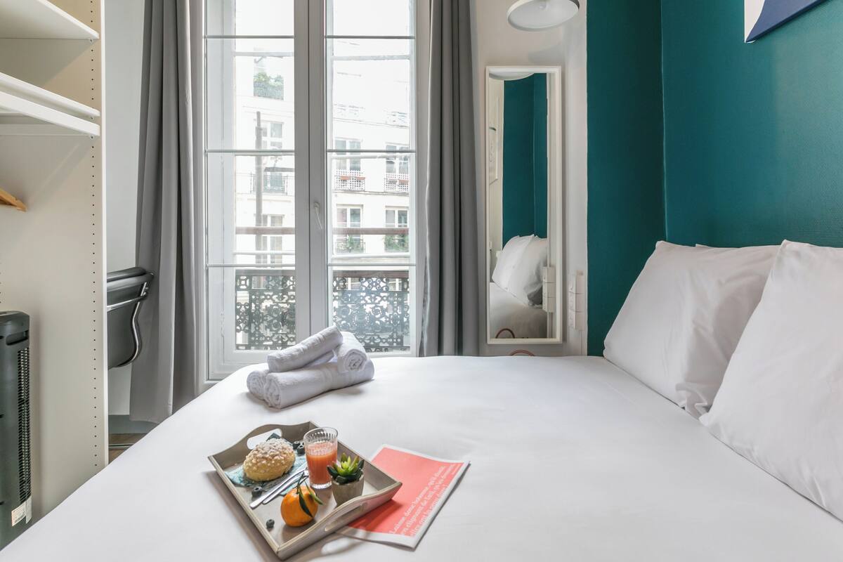 Une chambre d’hôtel avec un lit, un plateau avec de la nourriture et des boissons, et une fenêtre donnant sur des immeubles.