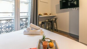 A hotel room with a bed, a tray with a drink, fruit, and towels. There is a TV mounted on the wall and a balcony with a railing. - WS Louvre - Richelieu (Paris)