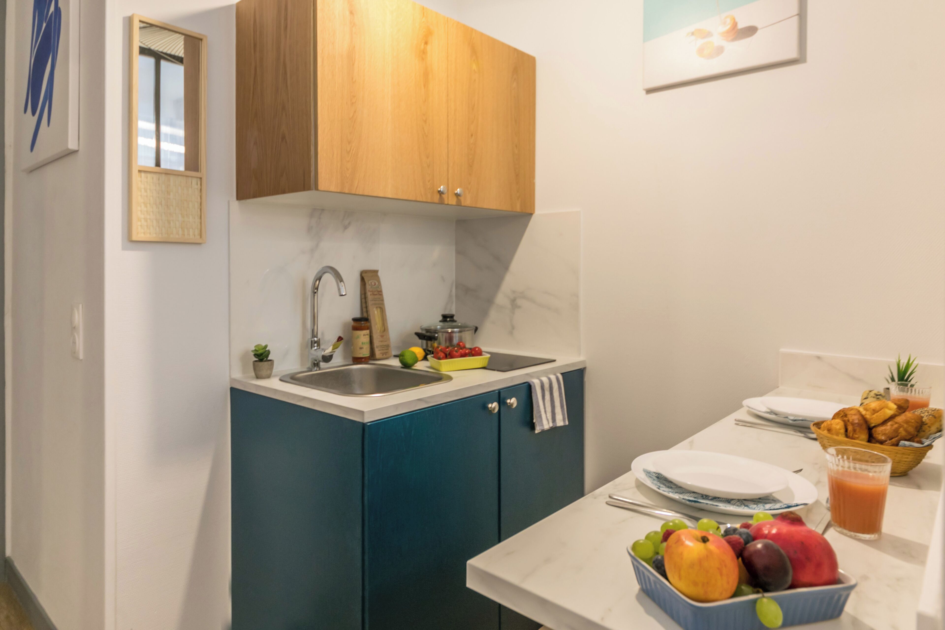 A modern kitchen with blue cabinets, a marble countertop, a sink, and a breakfast setup including fruits, a glass of orange juice, and a basket of pastries.