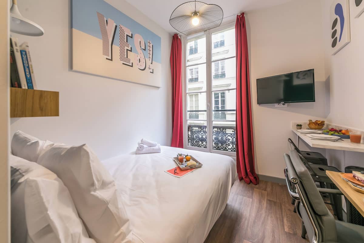 Une chambre d’hôtel avec un lit, un bureau où le petit-déjeuner est servi, une télévision et une fenêtre avec des rideaux rouges.