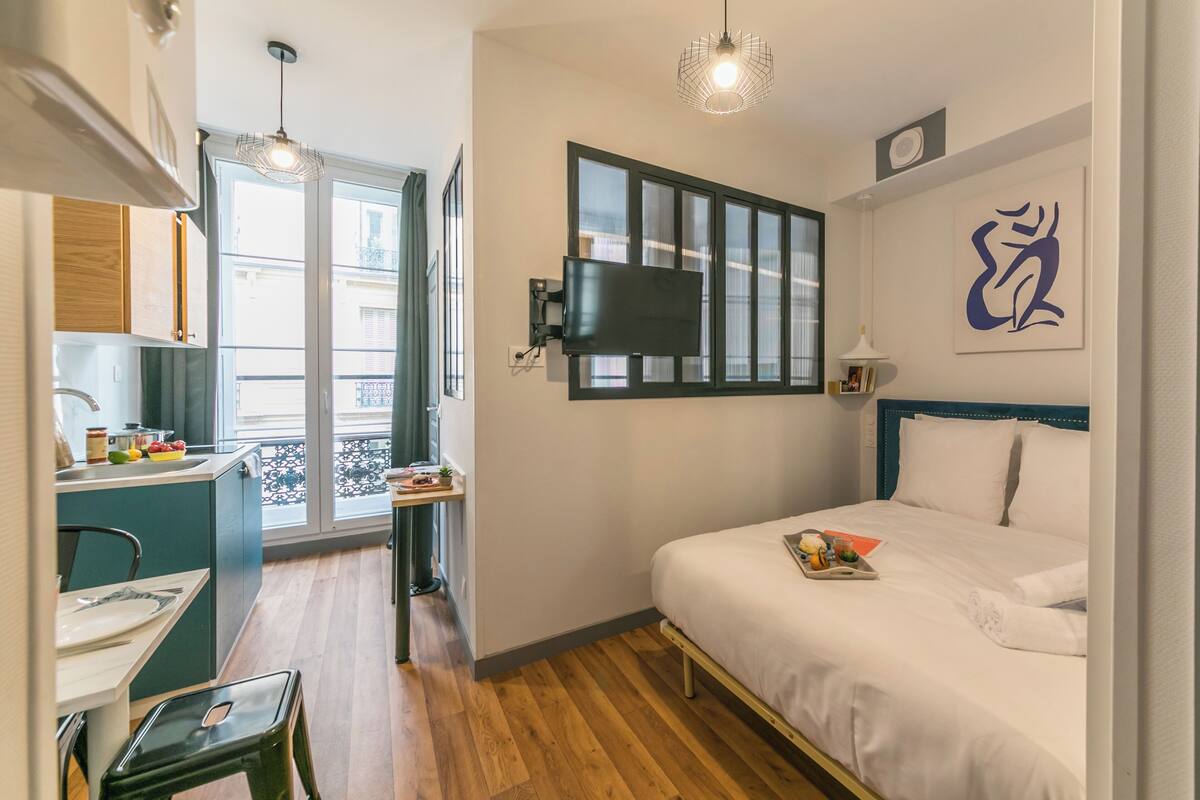 Une chambre d’hôtel compacte comprenant un lit, un bureau, une chaise, une fenêtre avec stores, un téléviseur fixé au mur et un petit balcon.