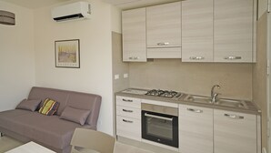 Une cuisine compacte équipée d’une cuisinière à gaz, d’un four et d’un évier encastré. On y trouve un canapé beige avec un coussin et un tableau encadré au mur.