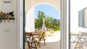 Eine geöffnete Tür, die auf eine Terrasse mit Tisch und Stühlen hinausführt. Eine Schlüsselablage hängt an der Wand, und man sieht Bäume sowie einen klaren Himmel.