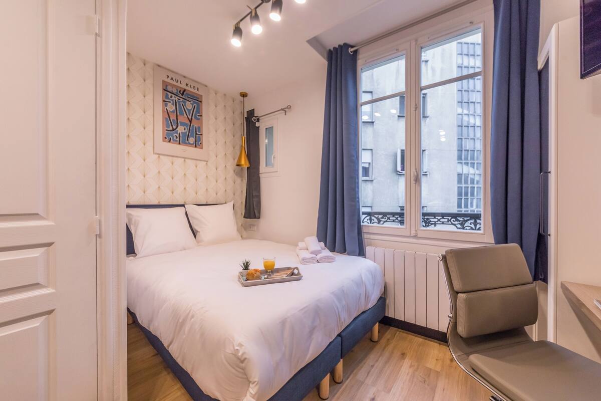 A neatly made bed with a tray of breakfast items, a window with curtains, and a view of the city.