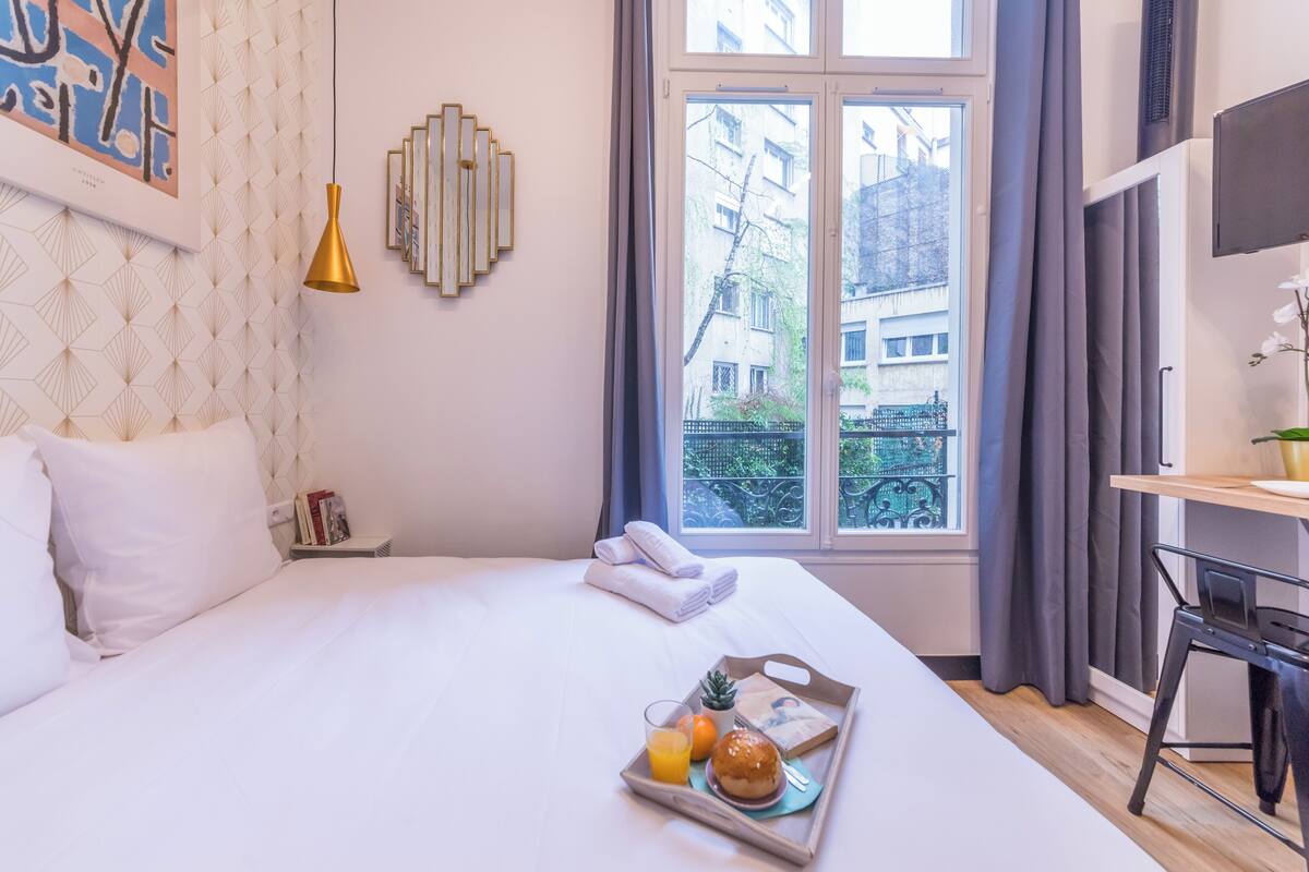 A hotel room with a large bed, a desk, a window with curtains, and a tray with breakfast items.