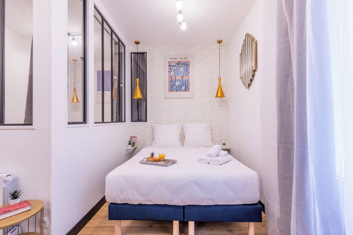 A modern hotel room with a large bed, a bedside table with a tray of fruit and a glass of orange juice, a small table with a book and a plant, and a large mirror on the left wall.