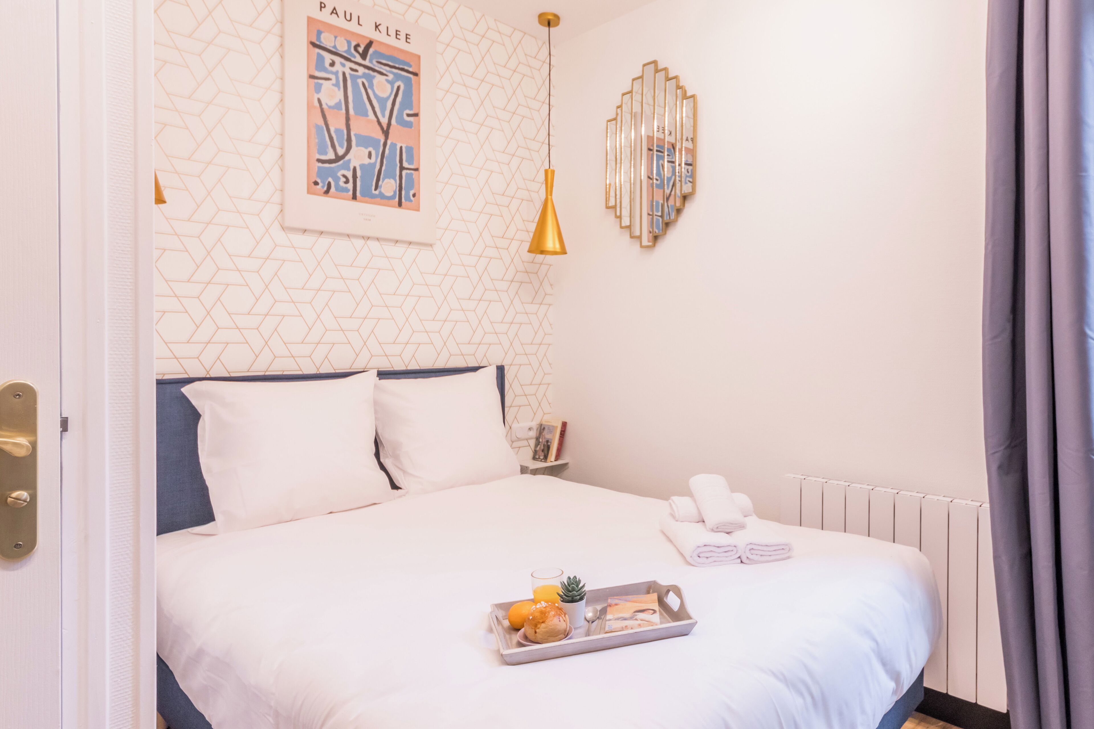 Una cama bien hecha con ropa blanca, una bandeja con alimentos para el desayuno y una pared con patrones geométricos y una obra de arte enmarcada.